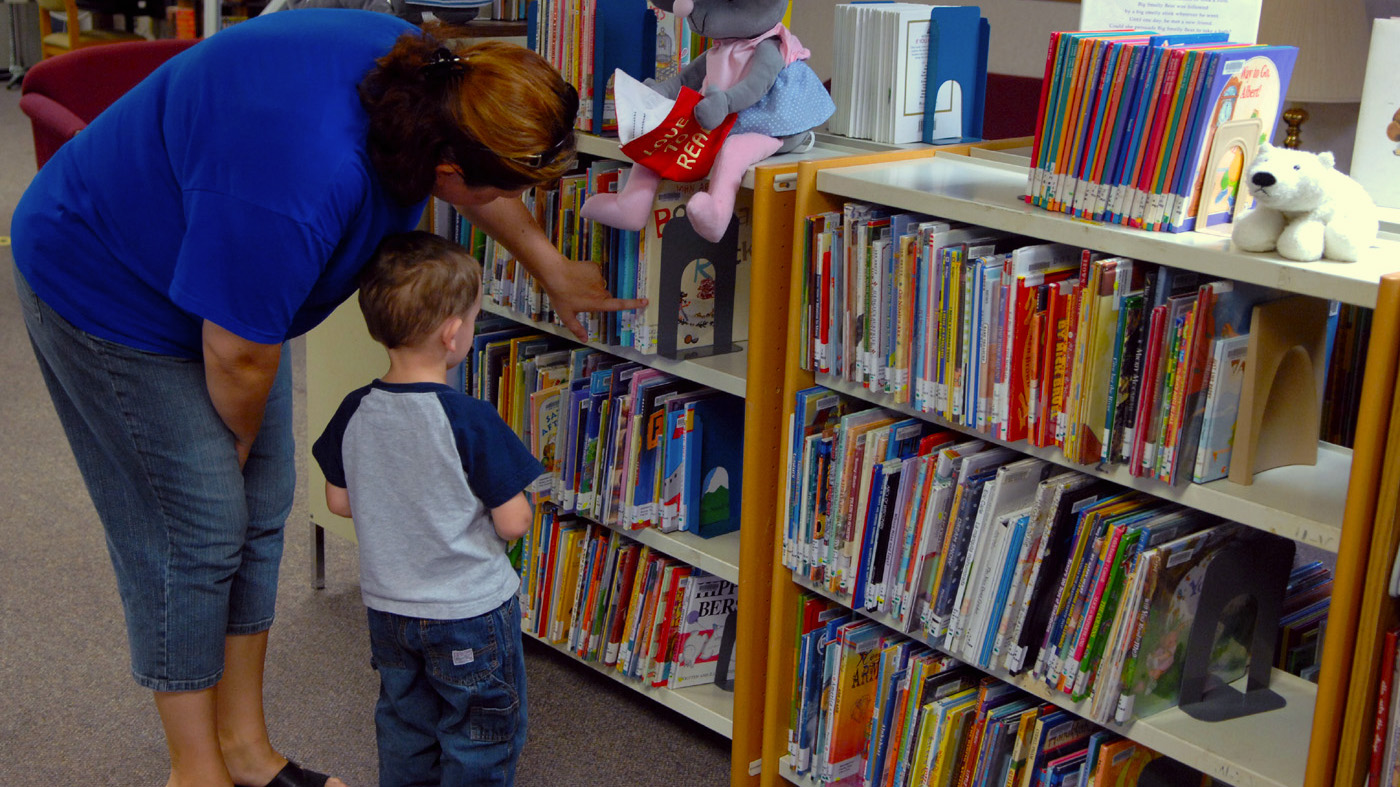 Librarian and small child search on a wooden library shelf containing lots of children's books.