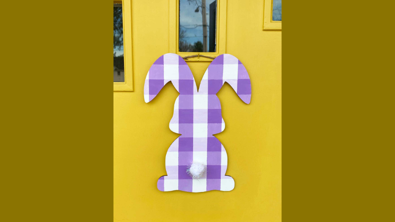 Wooden bunny painted in white and purple stripes hanging from a yellow door.